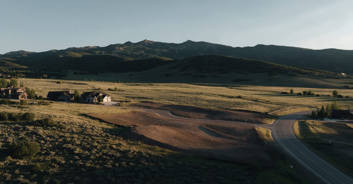 Golden-hour view over open sagebrush land at the edge of new homes near Park City, with graded lots and the Wasatch ridgeline beyond.